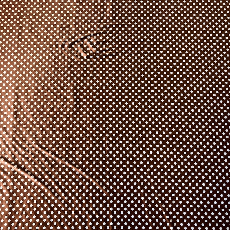 May include: Brown fabric with a white polka dot pattern. The fabric has a slight sheen and is folded in places, creating shadows and variations in the color. The polka dots are evenly spaced.