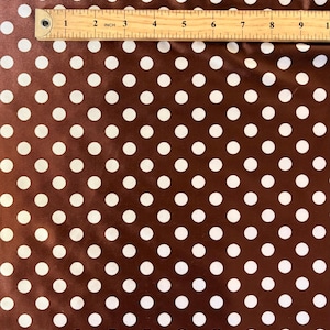May include: Brown fabric with white polka dots. A wooden ruler is placed across the top of the fabric, showing measurements in inches. The polka dots are evenly spaced and create a classic pattern.