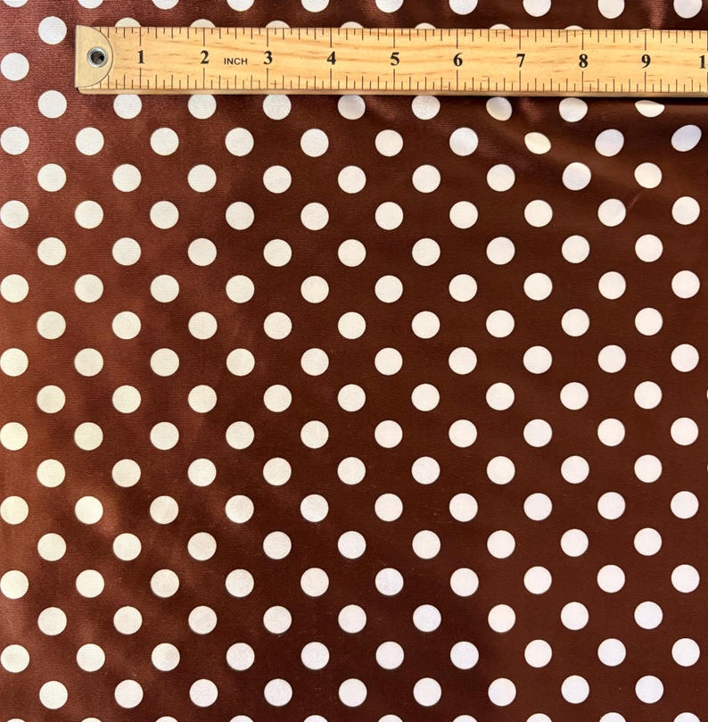 May include: Brown fabric with white polka dots. A wooden ruler is placed across the top of the fabric, showing measurements in inches. The polka dots are evenly spaced and create a classic pattern.