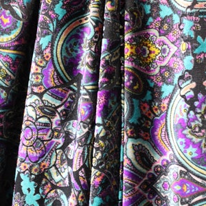 Op de afbeelding: Close-up van een stof met een paisley-patroon in tinten turquoise, paars, roze en geel op een zwarte achtergrond.