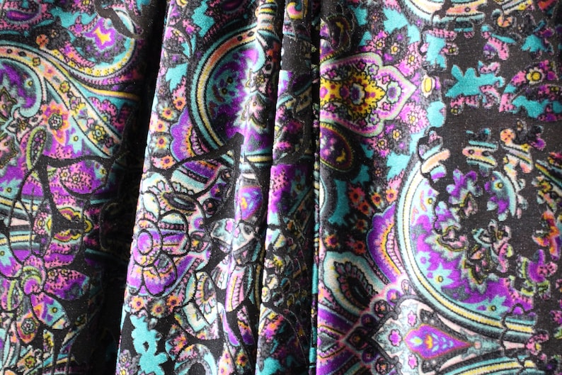 Op de afbeelding: Close-up van een stof met een paisley-patroon in tinten turquoise, paars, roze en geel op een zwarte achtergrond.