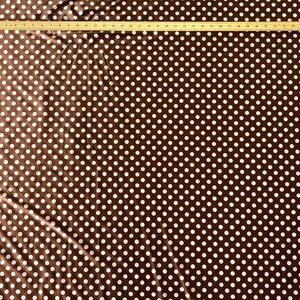 May include: Brown fabric with a white polka dot pattern. A wooden ruler is placed across the top of the fabric. The fabric is a dark brown color with evenly spaced white polka dots.