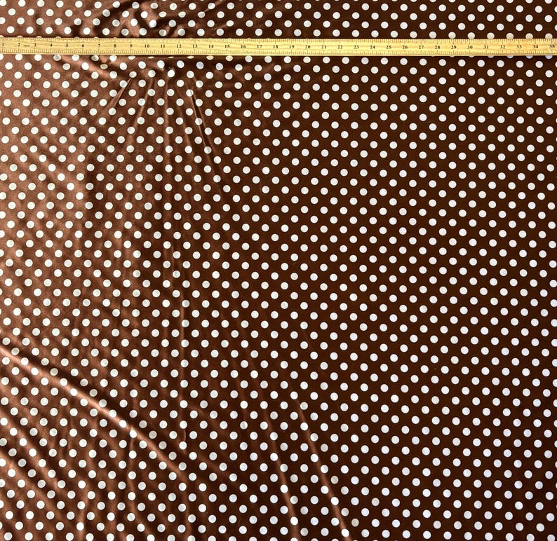 May include: Brown fabric with a white polka dot pattern. A wooden ruler is placed across the top of the fabric. The fabric is a dark brown color with evenly spaced white polka dots.