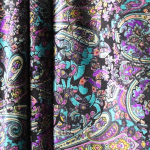 Op de afbeelding: Een close-up van een stof met een paisley-patroon in tinten turquoise, paars, roze en wit op een zwarte achtergrond.