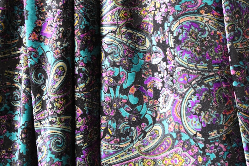 Op de afbeelding: Een close-up van een stof met een paisley-patroon in tinten turquoise, paars, roze en wit op een zwarte achtergrond.