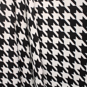 May include: Black and white houndstooth fabric featuring a repeating pattern of dog tooth shapes. The fabric is draped, showing folds and texture. The classic design is suitable for clothing and home decor.