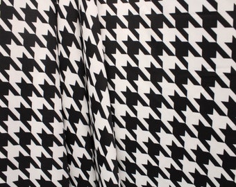 May include: Black and white houndstooth fabric with a repeating pattern of stylized dog tooth shapes. The fabric is draped, showing folds and texture. The classic design is suitable for apparel and home decor.
