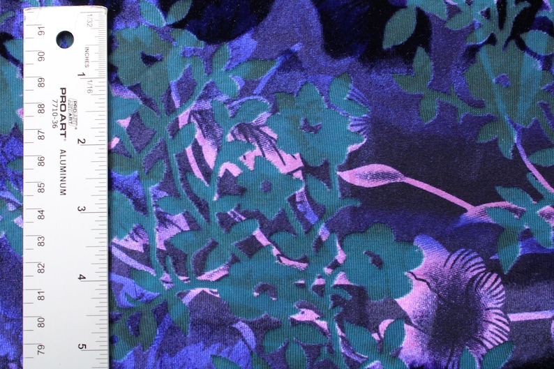 May include: A close-up of a teal, purple, and pink floral fabric with a textured, embossed design. The fabric is measured with a ruler in inches.