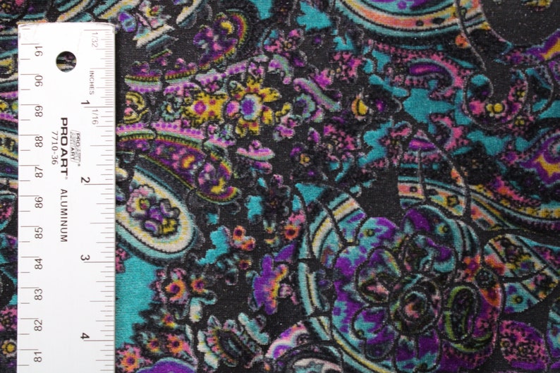 Op de afbeelding: Een close-up van een zwarte stof met een paisley-patroon in tinten roze, paars, turquoise en geel. Een liniaal is zichtbaar aan de linkerkant van de afbeelding.