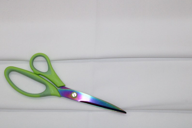 May include: A pair of green handled scissors with a rainbow iridescent blade on a white surface.