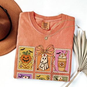 Op de afbeelding: Koraalrood Comfort Colors T-shirt met een Halloween-thema. Het ontwerp bevat postzegelachtige afbeeldingen van een pompoen, een spook en een koffiekopje, allemaal met medische thema's. Een gouden ketting en een bruine hoed zijn ook zichtbaar.