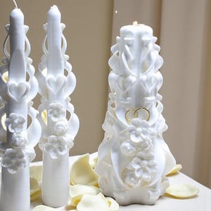 May include: White carved candles with floral and heart designs. One candle features two gold rings. The candles are on a white surface with scattered petals. The candles are a decorative item.