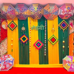 May include: A colourful display featuring umbrellas, diamond-shaped decorations, and vertical stripes of yellow and green fabric. The word "Mehndi" is displayed in green script. The scene is set against a red floor and floral accents.