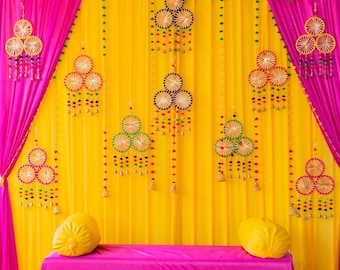 May include: A colourful backdrop for a wedding or event, with yellow and pink fabrics. Decorative circular ornaments with colourful accents hang from the top. The scene includes a pink table and yellow cushions, creating a festive atmosphere.