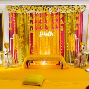 May include: A colourful stage decorated with yellow, pink, and green drapes, adorned with a floral arrangement and a neon sign reading "Dholki". The scene is enhanced with decorative tassels and fairy lights, creating a celebratory ambiance.