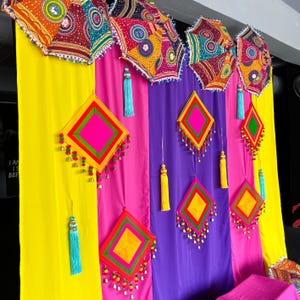 May include: A colourful display with patterned umbrellas and diamond-shaped decorations. The umbrellas feature intricate patterns in red, orange, and blue. The diamond shapes are in pink, yellow, and green, with tassels and small bells. The backdrop is striped with yellow, pink, and purple.