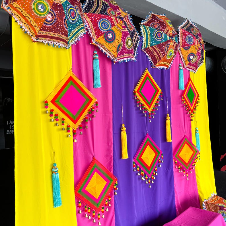 May include: A colourful display with patterned umbrellas and diamond-shaped decorations. The umbrellas feature intricate patterns in red, orange, and blue. The diamond shapes are in pink, yellow, and green, with tassels and small bells. The backdrop is striped with yellow, pink, and purple.
