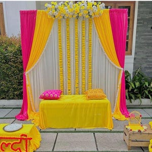 May include: A bright, colourful wedding or event backdrop with pink and yellow draped fabric, white curtains, and garlands of yellow flowers. A yellow table is adorned with pink and yellow cushions. A small table with the word "Haldi" is in the foreground.
