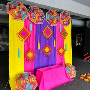 May include: A colourful display with patterned umbrellas, diamond-shaped decorations, and a draped table. The umbrellas and decorations feature intricate patterns with red, yellow, and blue accents. The backdrop includes yellow, pink, and purple fabric.