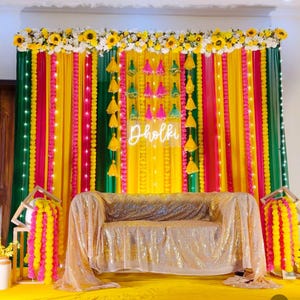 以下が含まれることがあります： 黄色、緑、ピンクのドレープ生地を使用した、活気のあるイベントの背景。 「Dholki」の文字が白いネオンで照らされています。 ひまわり、タッセル、金色のスパンコールソファで飾られたシーンです。