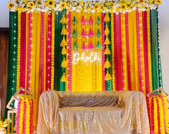 May include: A colourful event backdrop with yellow, green, and pink draped fabric. The word "Dholki" is illuminated in white neon. The scene is decorated with sunflowers, tassels, and a gold sequined sofa.
