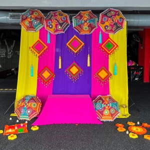 May include: A colourful display with decorative umbrellas, diamond-shaped ornaments, and draped fabric in yellow, purple, and pink. The umbrellas and ornaments have intricate patterns and embellishments, creating a festive atmosphere.