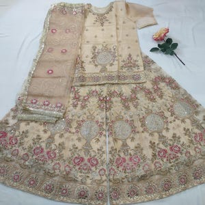 Puede incluir: Conjunto de ropa formal bordada de color crema. El conjunto incluye una parte superior de manga corta, pantalones de pernera ancha y una bufanda transparente. Patrones florales y geométricos con acentos rosas y dorados adornan la tela. Una rosa descansa cerca.