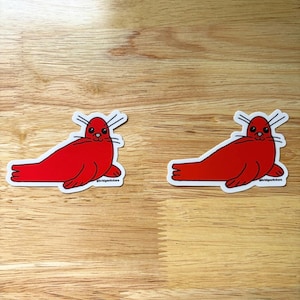 May include: Two red seal stickers with white borders are shown on a wooden surface. Each sticker depicts a cartoon seal in a resting pose, with white whiskers and a small black eye. The stickers are identical, with the text "@bridgestickers" at the bottom.