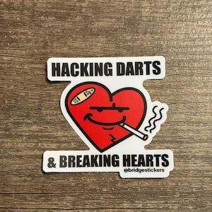 May include: A sticker with a red heart design, featuring a bandage, a cigarette, and a smiling face. The sticker has the text "HACKING DARTS & BREAKING HEARTS" in black, with the @bridgestickers logo.