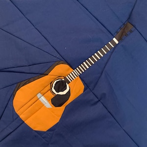 Könnte beinhalten: Ein orangefarbenes und braunes Akustikgitarren-Design auf einem dunkelblauen Hintergrund. Die Gitarre hat ein schwarzes Schallloch und weiße Bundmarkierungen. Das Bild ist eine Nahaufnahme eines Quilt- oder Stoffdesigns.