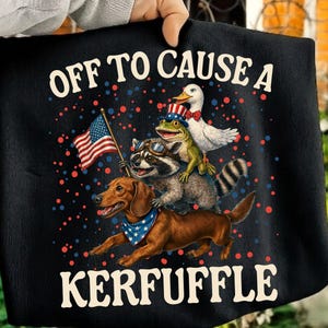 Peut inclure: Un t-shirt noir avec une illustration représentant un teckel, un raton laveur et un canard. Le teckel porte un bandana étoilé, le raton laveur des lunettes de protection et le canard un haut-de-forme patriotique. Texte : OFF TO CAUSE A KERFUFFLE.