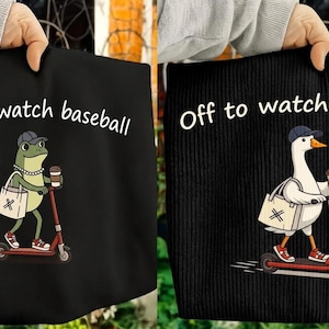 Peut inclure: Sac fourre-tout noir avec le texte "Off to watch baseball". Un sac présente une grenouille sur un scooter, l'autre une oie. Les deux personnages portent des chapeaux et portent du café et des sacs fourre-tout. La grenouille et l'oie font du scooter rouge.