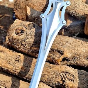 May include: A silver-colored sword with a black handle rests on a pile of wooden logs. The sword has a decorative hilt with rounded accents. The blade is long and pointed, and the handle is textured.
