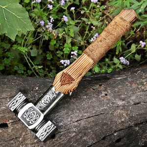 May include: A handcrafted wooden hammer with a metal head, featuring intricate Celtic knot designs. The handle is wrapped in twine for grip and has a carved triangular emblem. The hammer rests on a weathered wooden surface, with greenery in the background.