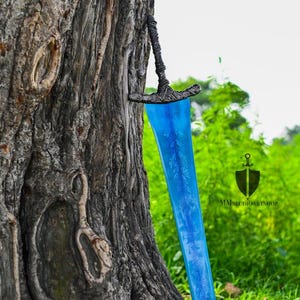 May include: A fantasy sword with a blue blade and ornate hilt is mounted on a tree trunk. The blade has a floral design. The hilt is dark gray and the blade is a vibrant blue. The sword is displayed outdoors.
