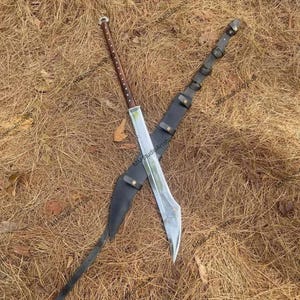 May include: A large, silver-bladed sword with a brown wooden handle and a black leather sheath. The sword is crossed over the sheath, resting on a bed of dry, brown grass.