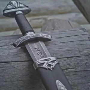 May include: A detailed image of a Viking sword. The sword features a dark wooden handle, a silver-colored guard with intricate designs, and a blade with etched text. The scabbard is dark with silver accents. The sword is resting on a wooden surface.