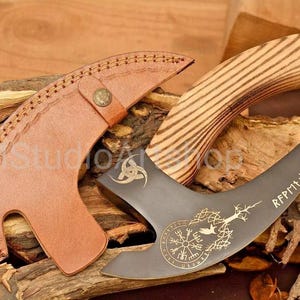May include: A handcrafted axe with a wooden handle and a dark metal blade. The blade features engraved symbols and text, with a leather sheath. The handle is made of light-colored wood with dark stripes.