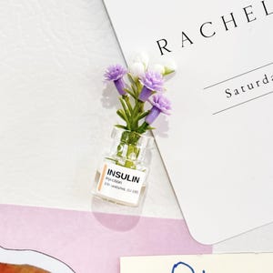 Könnte beinhalten: Ein kleines Glasfläschchen mit der Aufschrift "INSULIN injection 100 units/mL (U-100)" enthält einen kleinen Strauß lila und weißer Blumen. Das Fläschchen steht vor einem weißen Hintergrund mit einer Karte mit der Aufschrift "RACHEL" und "Saturday".