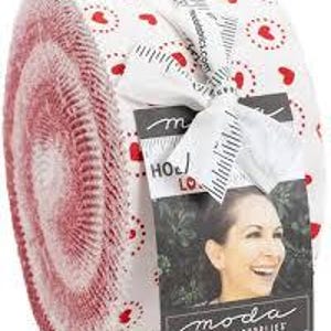 May include: A rolled fabric bundle with a red and white heart design. The fabric is secured with a white ribbon and a tag that reads "HOLIDAY LOVE". The bundle is labelled "MODA FABRICS - SUPPLIES".
