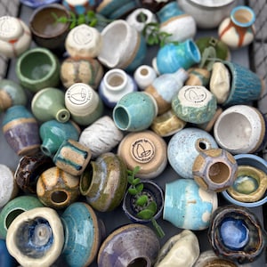Puede incluir: Una colección de pequeñas macetas y recipientes de cerámica hechos a mano en varias formas y tamaños. La cerámica presenta una gama de colores, incluyendo azules, verdes, marrones y blancos, con algunas piezas que muestran superficies texturizadas y diseños únicos. Algunos tienen el texto "art shed" y "Trina".