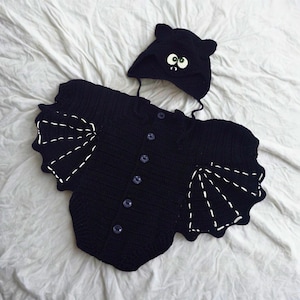 Pode incluir: Um conjunto de bebê de crochê preto com asas de morcego e um chapéu combinando. O traje tem fechos de botão na frente e detalhes de costura brancos nas asas. O chapéu apresenta orelhas de morcego e olhos de desenho animado.
