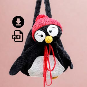 Pode incluir: Uma mochila de pinguim de crochê em preto e branco com um chapéu vermelho e um bico amarelo. O pinguim tem olhos brancos grandes e um cordão vermelho. A mochila é feita à mão e tem um ícone de download PDF.