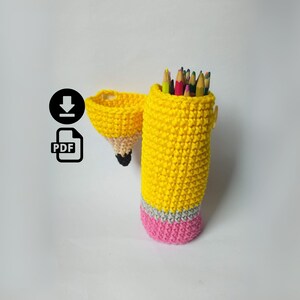 Pode incluir: Um porta-lápis de crochê amarelo feito à mão em forma de lápis, com base cinza e rosa. Contém lápis de cor. Um porta-lápis menor e combinando está anexado.
