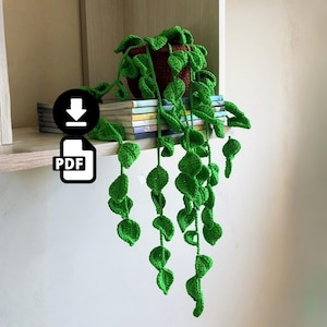 Pode incluir: Uma planta suspensa de crochê com folhas verde-vibrantes caindo de um vaso marrom escuro. A planta está sobre uma pilha de livros finos. Um ícone circular preto com uma seta branca para baixo e um ícone de documento PDF são visíveis.