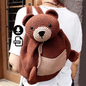 Pode incluir: Uma mochila de urso de crochê marrom com focinho e bolso bege. A mochila tem alças ajustáveis e fecho de cordão. O rosto do urso apresenta olhos pretos e nariz preto.