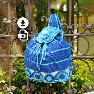 Pode incluir: Mochila de crochê azul feita à mão com fecho de cordão e aba. A bolsa apresenta um padrão floral detalhado na parte inferior e na aba, com detalhes em azul claro. A bolsa é exibida ao ar livre, pendurada em um portão de metal decorativo.
