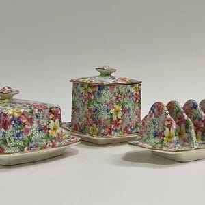 May include: A floral-patterned ceramic butter dish with a lid, a square lidded container, and a toast rack. The pieces feature a light green background with pink, yellow, and blue flowers. The edges are trimmed in gold.
