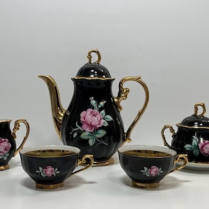 May include: A black porcelain tea set with gold trim, including a teapot, creamer, sugar bowl, and two teacups. Each item is adorned with a pink rose and green leaves. The set is designed for serving tea.