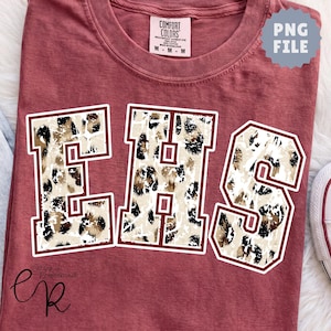 May include: A brick red t-shirt featuring the letters EHS in a large varsity font. The letters are filled with a tan, brown, and black leopard print pattern and outlined in white and dark red. A Comfort Colors brand label is visible at the neck.